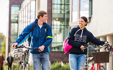 Two students walk side by side wheeling their bicycles through UCD campus.